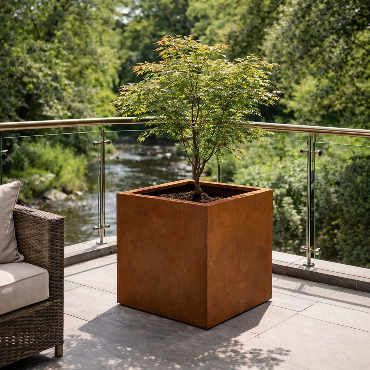Corten steel cube planter with maple tree on modern terrace overlooking stream – designer garden planter UK